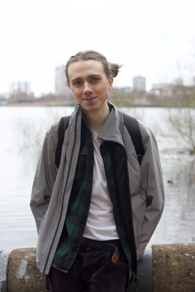 Portrait of Isaac, white, non binary person in a cycling jacket and space buns at Edgbaston Reservoir.