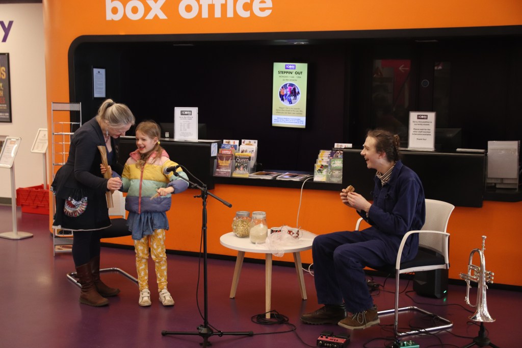Photo of Isaac with a parent and child - with household items and microphone on a makeshift stage. Everyone's smiling.